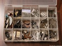 Top view of open plastic organizer with multiple compartments filled with assorted silver-tone jewelry crafting charms, pendants, and findings.