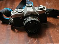 Front angled view of Minolta XG-A camera with attached 50mm lens and strap lying on wooden surface.