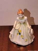 Front view of Royal Doulton March Pretty Ladies porcelain figurine wearing a white dress with yellow flowers and holding a hat.