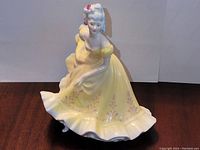 Front angled view of the Ninette figurine showing detailed face, floral hair decoration, and flowing yellow gown with purple floral patterns.