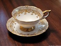 Teacup and saucer set displayed on wood table showing detailed gold floral pattern and gold accented handle.