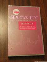 Front cover of the sealed DVD box set showing burgundy background with city skyline outline and title 'Sex and the City The Complete Series' with HBO logo and sticker detailing contents including 6 Seasons, 94 Episodes, 20 Discs.