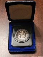 Top view of commemorative silver dollar coin in protective casing, positioned inside blue velvet-lined area of black presentation box with beige interior cushioning on the lid.