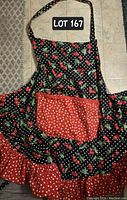 Front side of the apron showing cherry motif on dark background with white polka dots, red polka dot ruffled bottom and matching pocket. Black and white polka dot neck strap visible.