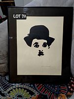 Framed black and white minimalist painting of Charlie Chaplin face with bowler hat, displayed against fabric background.