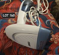 Side angle of white and blue handheld Conair fabric steamer lying on patterned fabric background, with power cord tangled behind.