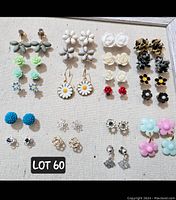 Photo showing 22 pairs of floral stud and small dangly earrings laid out in rows on a beige background inside a wooden frame, showcasing various colors and designs including white, cream, red, blue, green, pink, and tortoiseshell tones.