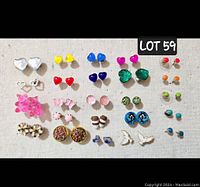 Top-down photo displaying 24 pairs of various colorful fun stud earrings arranged in rows on a beige surface.