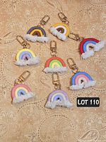 Photo showing 8 handmade macrame rainbow keychains arranged on a beige patterned fabric backdrop. Each keychain has a gold metal swivel lobster clasp and white fringed tassel clouds.