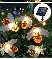 Close up of five illuminated honey bee-shaped LED lights at night among flowers, showing amber and clear plastic design with glowing light inside.
