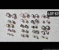 Photo displays 20 pairs of rhinestone stud earrings in various colors and designs arranged in rows on a beige surface.