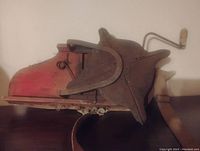 Side view of antique hand crank seed spreader showing red metal body, wooden crank handle, and star-shaped spreader mechanism.