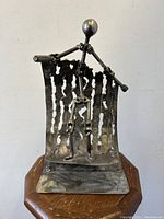 Front view of the brutalist metal sculpture showing the surfing figure standing on the curved sheet metal wave with a perforated texture, mounted on a flat metal base.