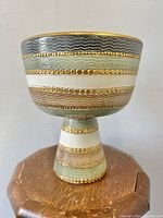 Side view of chalice form ceramic bowl on wooden pedestal, showing textured bands of gray, green, white and gold beadwork