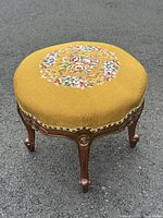 Full view of round wooden footstool with needlepoint floral tapestry upholstery on top and curved carved legs in wood brown finish.
