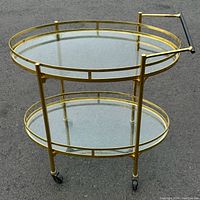 Side view showing two oval glass shelves with solid brass frame, slight rust visible on frame edges, black handlebar and caster wheels.