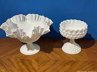 Front view of the large ruffled compote with hobnail texture and the smaller hobnail goblet bowl set on a wooden surface against a blue background.