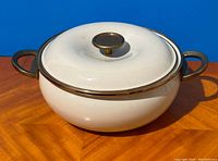 Front angle view showing off white enamel pan with lid, brass-tone handles and knob, on wood surface with blue background.