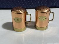 Front view of copper salt and pepper shakers showing brass plates with 'S' and 'P' markings, brass handles and tops with multiple holes.