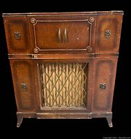 Front view of vintage Electrohome stereo cabinet showing wood finish, central speaker grille with fabric and diamond lattice overlay, side compartments with metal ring handles and upper double doors with handles.