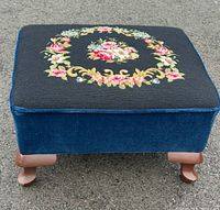 Front view of the vintage footstool showing blue velvet sides and wooden legs, with a slight view of needlepoint embroidery on top.