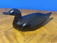 Front and side view of the hand carved dark wood duck on a wood surface with blue background