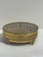 Full view of round vintage brass and glass jewelry trinket box showing ornate filigree work and decorative claw feet.