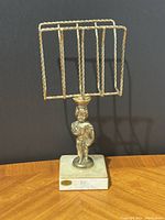 Full view of the vintage brass napkin or letter holder with Roman figure standing on white marble base, shown against a black background on a wood surface.