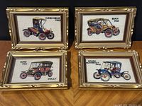 Photo showing the four vintage framed needlepoint artworks depicting Oldsmobile 1902, Buick 1910, Ford 1911, and Brush Runabout 1912 cars in gold decorative frames.