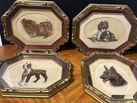 Four framed needlepoint artworks featuring dogs, arranged on wooden surface showing detailed embroidery and frames.