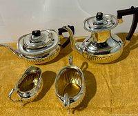 Set of four silver plated pieces including two teapots, a cream jug, and a sugar bowl arranged on a yellow cloth.