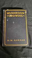 Front cover of the antique book titled 'Mushroom Growing' by B.M. Duggar, black cover with yellow text, some marks visible.