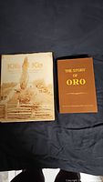 Two hardcover books laid side by side on black cloth background; 'The Story of Oro' brown cover and 'Kith 'N Kin' illustrated dust jacket
