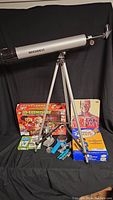 Full lot image showing Bushnell telescope on tripod, chemistry kit box, microscope, magnifying glass, books and flash cards arranged on black cloth background.
