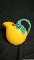 Side view of ceramic pitcher shaped like an orange with green leafy handle against black background.