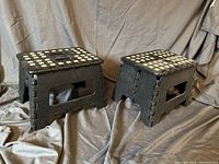 Two black plastic folding step stools with white squares on the top surface, positioned side by side on a grey cloth background.