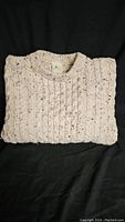Folded cream Aran knit sweater showing knit patterns and crew neckline on black background.