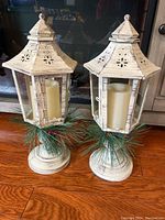 Pair of white distressed lanterns showing detail of hinged glass door, battery candle inside, and greenery base