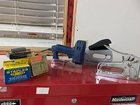 All three staple guns and assorted boxes of staples arranged on a red surface under a window showing the set contents.