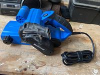 Photo of Mastercraft blue and black electric belt sander on wooden surface, showing side profile and cord wrapped.