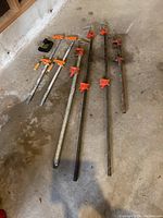 Overview of all seven clamps laid out on concrete floor showing two smaller metal bar clamps with orange handles and wooden grips, one black C-clamp, and four metal bar clamps of various sizes with red tightening mechanisms.
