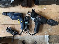 Photo of both the Mastercraft electric drill on the left and the Black & Decker heat gun on the right placed on a wooden surface showing condition and cord arrangements.