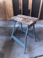 Front angle view of folding work bench showing metal gray legs with crossbar support and well-worn wooden top with holes.