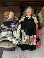 Two vintage dolls in black and white traditional dresses with lace and floral detailing, one braided hair