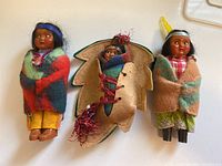 Photo showing front view of all three vintage Skookum dolls, two wrapped in colorful felt shawls and one inside leaf-shaped felt carrying sack.