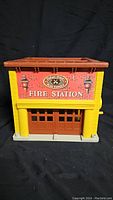 Front view of the vintage Fisher Price Fire Station showing red and yellow plastic building with 'Fire Station' label and garage doors.