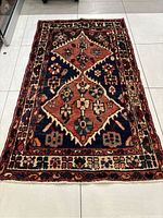 Full view of the Bakhtiari rug showing geometric and floral patterns in natural colors of red, navy blue, cream, and black on tiled floor.