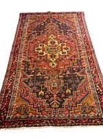 Full view of the Persian rug showing geometric central medallion and border design in red, black, ochre, and yellow tones with fringed edges.