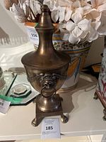 Photo showing the vintage brass urn with lid adorned with lion head motifs holding rings and claw feet base. The urn exhibits an aged patina.
