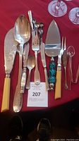 Various classic utensils laid out flat on red surface showing assortment of sizes and styles including fish knives, spoons, fork, serving tools with yellow resin and turquoise bead handles.